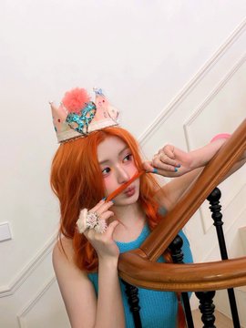 A young woman with orange hair wearing a colorful crown poses on a staircase, holding a wooden stick to her face.