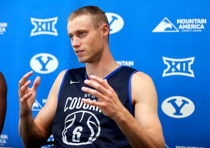 A basketball player wearing a navy blue BYU Cougars jersey (#6) speaking at a press conference, with BYU and Big 12 logos visible on the blue backdrop behind him.