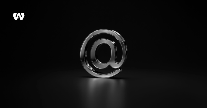 A glossy metallic @ symbol is centered on a dark black background, with a small white logo in the upper-left corner.