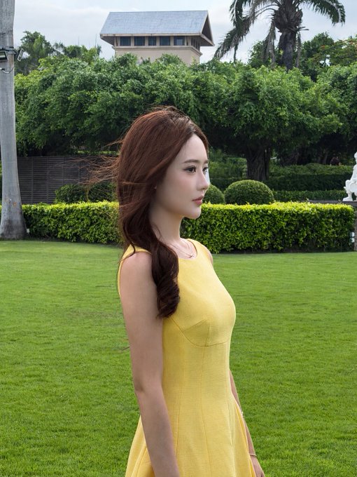 A young woman with long reddish-brown hair wearing a yellow sleeveless dress stands in profile on a manicured green lawn, with lush tropical hedges and a building visible in the background.