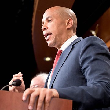 A bald man in a navy blue suit and red tie speaks passionately at a podium while gripping a microphone.