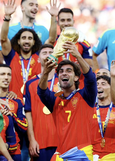The image shows the Spanish national football team celebrating with the FIFA World Cup trophy, with a player wearing jersey number 7 hoisting the gold trophy high while teammates, including those in the background, cheer and wear gold medals around their necks in their red and blue kits. The scene captures the joy of a World Cup victory, contextualizing the generous donation by an anonymous squad member to Spanish FA employees as a gesture of giving back following their triumphant success on the global stage.