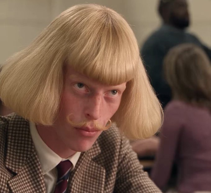 A man wearing a blonde bob wig and curled handlebar mustache stares intently forward, dressed in a houndstooth jacket and striped tie.