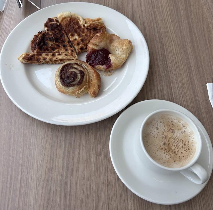 A white plate of assorted pastries including a cinnamon roll, Danish, and waffle pieces, served alongside a cup of coffee with foam on a wooden table.