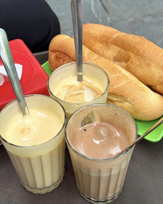 A close-up of three glasses containing pale yellow, creamy yellow, and chocolate-colored beverages with spoons, alongside a baguette on a green tray.