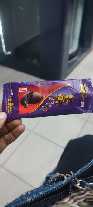 A hand holds a purple 'Hide & Seek Centre Filled Chocolate Coated' biscuit package with a chocolate cookie image visible.