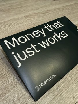 A sleek black PlasmaOne card rests on a wooden surface, featuring the tagline "Money that just works" in bold white lettering. The card represents the physical U card mentioned in the post, showcasing its minimalist design being delivered to members in the Asian region.