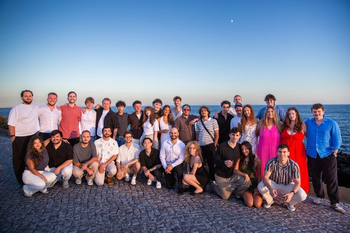 A large group photo of approximately thirty people posing together on a stone pathway near the ocean at sunset.