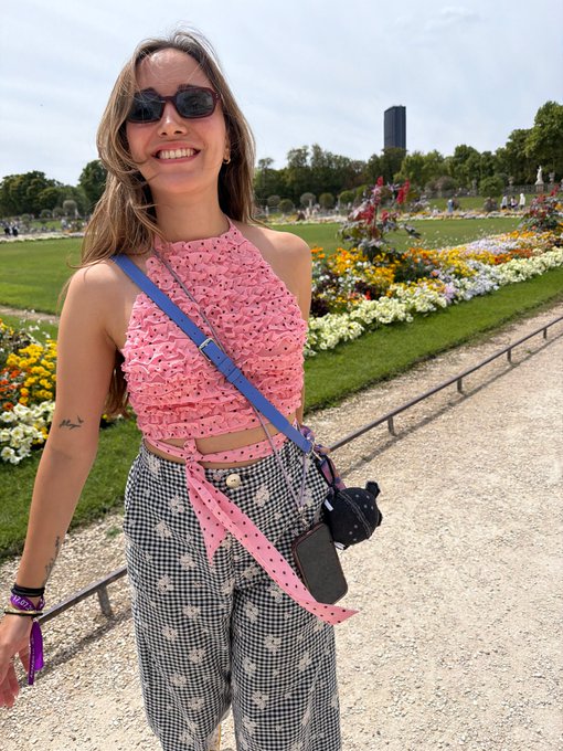 A smiling woman wearing sunglasses, a pink halter top, and houndstooth-patterned pants poses outdoors in a park with colorful flower beds and a distant tall structure visible in the background.