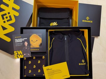 A Binance-branded merchandise box set containing a dark hoodie with yellow Binance logo, a yellow cap, and other branded items.