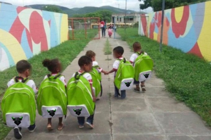 A group of small children wearing matching bright green backpacks with bull logos walk down a paved path lined with colorful painted walls.