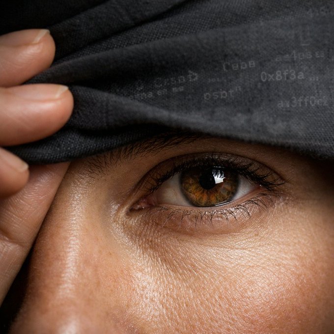 A close-up of a person lifting dark fabric to reveal one eye, with faint digital text and numbers, including “0x8f3a,” reflected on the material.