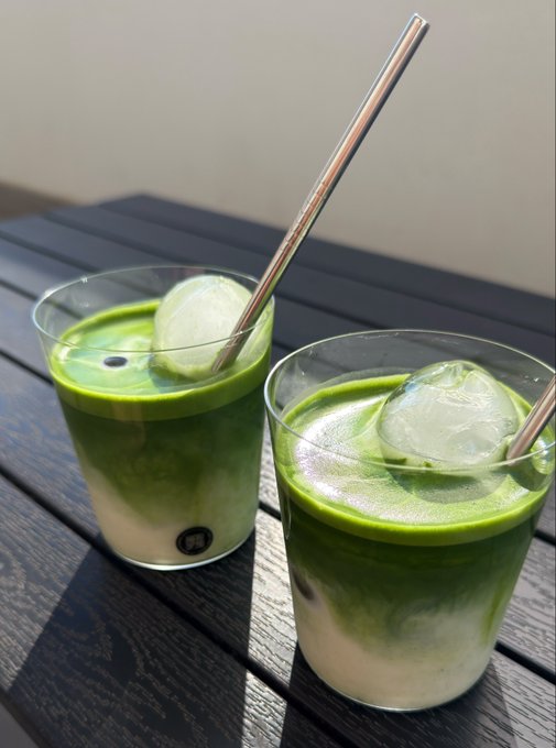 Two glasses of iced matcha latte with metal straws sit on a dark wooden slatted table in the sunlight.