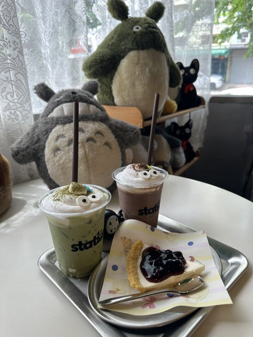 Two beverages from 'statio' cafe and a slice of cheesecake are served on a tray, with Totoro plush toys visible in the background.