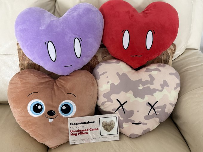 Four heart-shaped plush pillows with different faces (purple, red, brown with big eyes, and camo with X eyes) displayed on a couch with a card reading 'Congratulations! You won an Unreleased Camo Hug Pillow'.