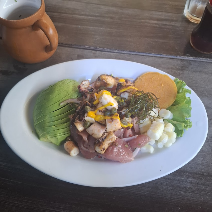 The image shows a white plate filled with food placed on a wooden table. The dish consists of various ingredients such as fish, avocado, and other vegetables. A cup can be seen next to the plate, possibly containing a beverage or condiment. The caption reads "This is my lunch today: Ceviche Paid with @useTria, as always", indicating that the person who ordered this meal has chosen to have ceviche for their lunch.