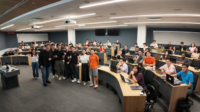A large group of people pose for a photo inside a modern lecture hall or classroom with curved desks and computer monitors.