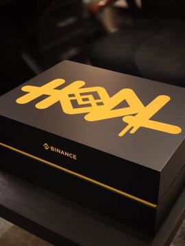 A sleek black Binance 9th anniversary VIP swag box with gold "X" lettering sits on a dark surface, representing the celebratory package given in honor of the exchange's milestone of 300M+ registered users.