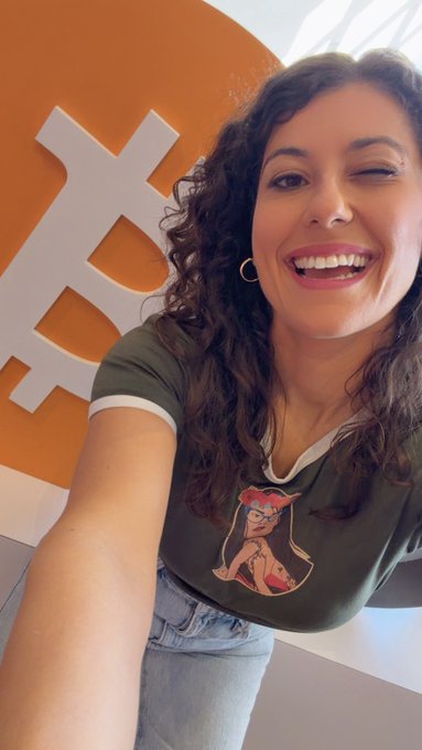 The image features a woman with curly hair wearing a green shirt, smiling at the camera while taking a selfie. The background of the photo includes an orange Bitcoin sign or logo, which adds a unique touch to the scene. The caption "gm I don’t care how low bitcoin is I’m still buying it see yall in 203o" suggests that the woman might be interested in cryptocurrency and wants to share her enthusiasm with others through this photo.