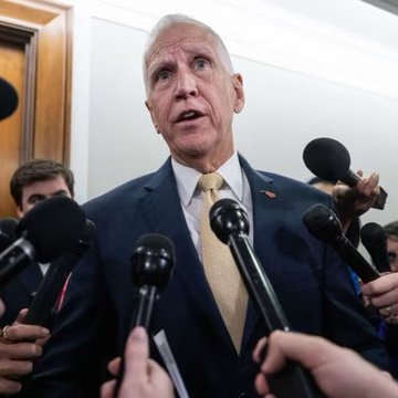 A man in a dark suit and gold tie speaks to a cluster of microphones held by reporters, appearing to address the press.