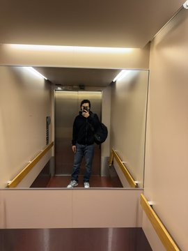 A man in dark clothing stands in an elevator, captured as a reflection in the mirrored elevator door.