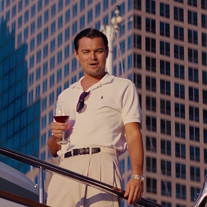 Leonardo DiCaprio stands on a boat wearing a white polo shirt and cream trousers, holding a glass of red wine with sunglasses tucked into his collar, set against a backdrop of skyscrapers.
