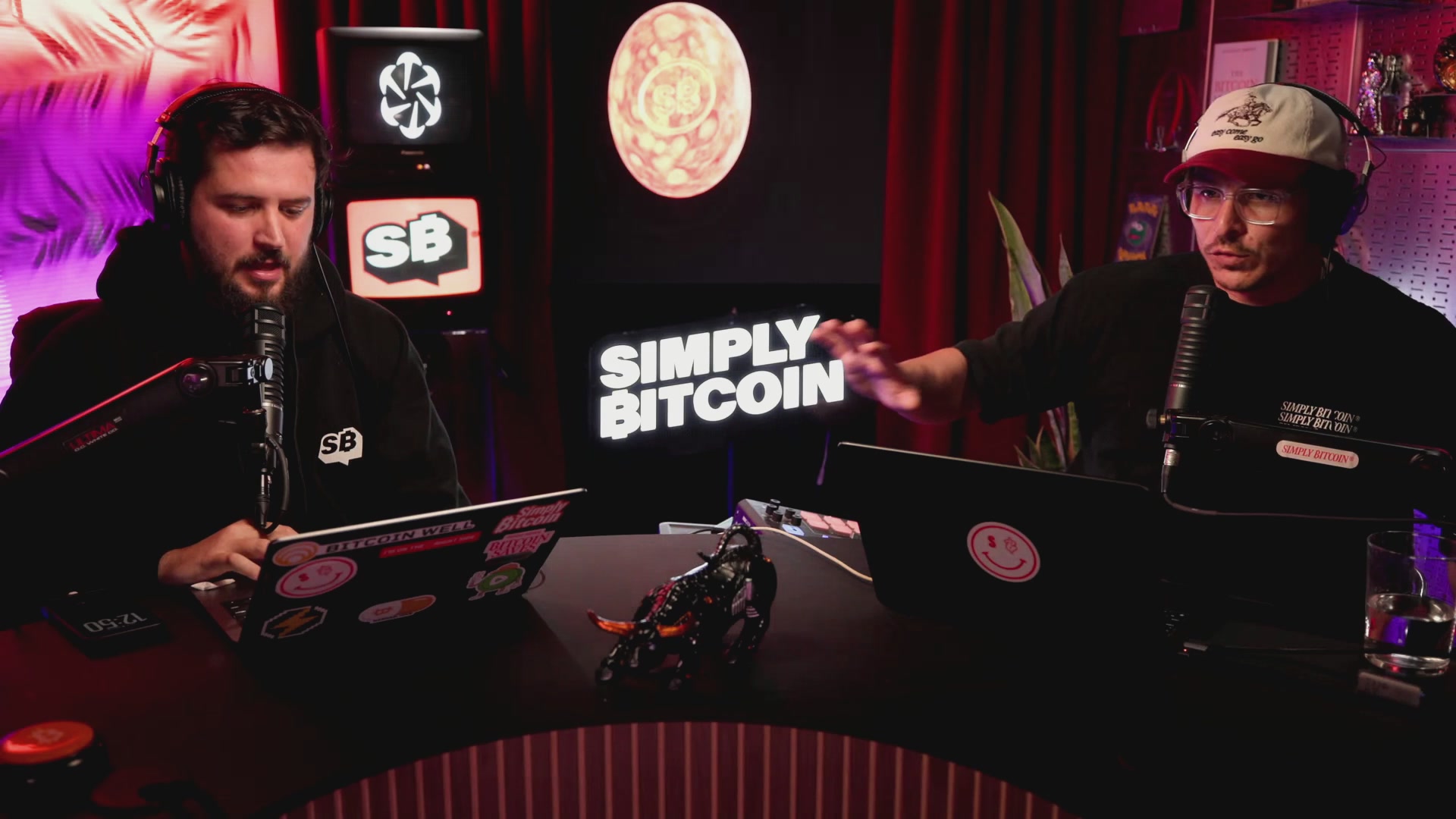 The image shows two men sitting at a table with laptops open, possibly working or discussing something related to cryptocurrency. The background features a red curtain and a sign that says "Simply Bitcoin" written in white letters.