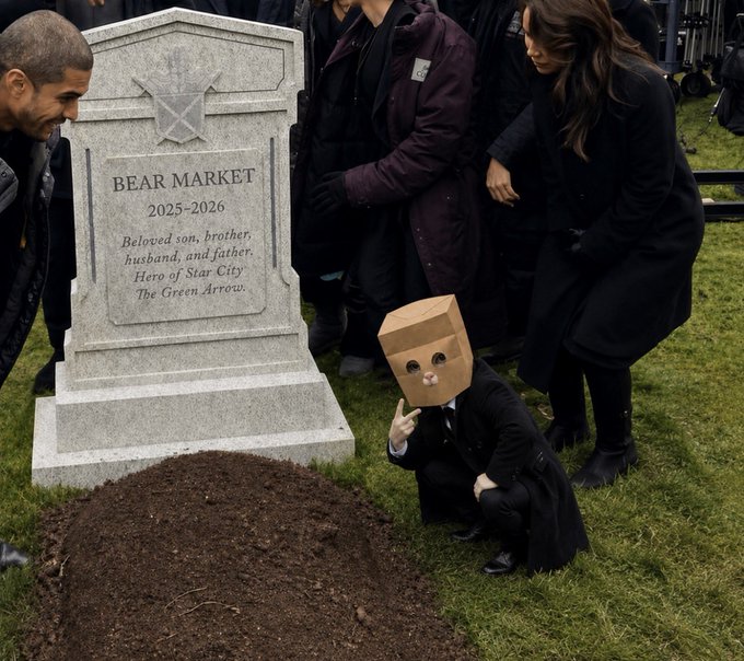 A funeral scene featuring a tombstone reading 'BEAR MARKET 2025-2026, Beloved son, brother, husband, and father. Hero of Star City. The Green Arrow.', with a figure in a black suit wearing a paper bag with cat face drawn on it kneeling at the grave.