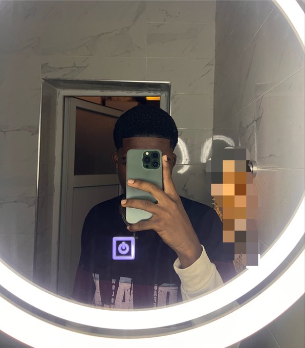 A mirror selfie of a young man taking a photo with a green iPhone, with another person partially visible in the background, reflected in a round illuminated bathroom mirror.