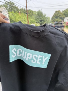 A hand holds up a black sweatshirt featuring a teal logo with the text "SCUPSEY" in bold letters. The image showcases a wearable representation of the Cupsey mascot mentioned in the post, worn as merchandise to represent the $Cupsey token.
