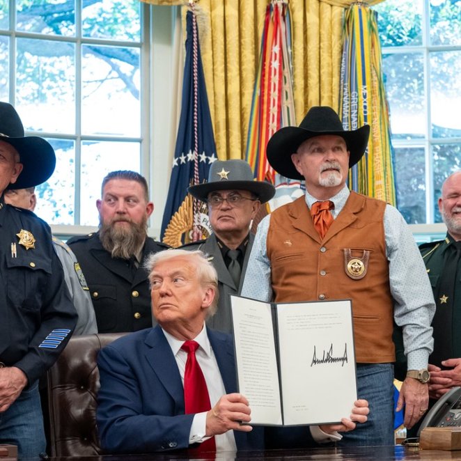 President Donald Trump holds up a signed document in the Oval Office, surrounded by members of the National Sheriffs' Association and other law enforcement officers in uniform and cowboy hats, as the group publicly endorses passage of the Clarity Act to support Bitcoin's growth in America.