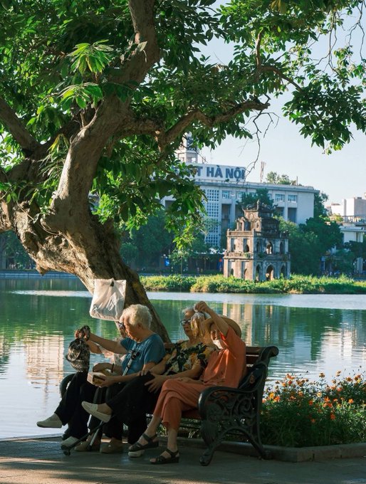 Three elderly people sit on a bench under a tree by Hoan Kiem Lake, with the iconic Turtle Tower visible across the water and a "HÀ NỘI" sign in the background. The post contrasts the carefree retired life depicted in the image—exercising and relaxing by the lake—with the stresses of hustling for paychecks and crypto investments.