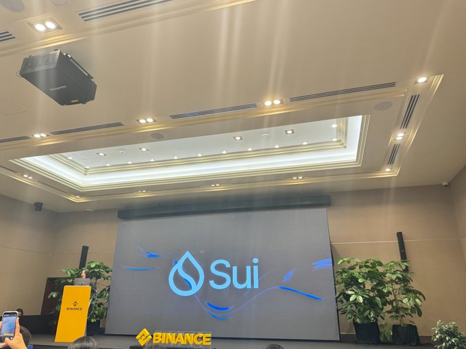 A conference room with a large screen displaying the 'Sui' logo and the 'BINANCE' logo on a podium.