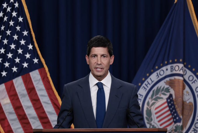 A man in a dark suit and blue tie speaking at a podium, with an American flag and the Board of Governors of the Federal Reserve System seal visible behind him.