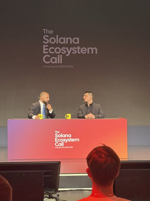 Two men sit at a desk on stage during 'The Solana Ecosystem Call' hosted by SOLBROTHERS, with the event title displayed on a large screen behind them and on the desk front.