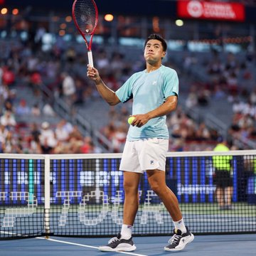 A tennis player in a teal shirt and white shorts standing on the court near the net, holding a red racket in his right hand and a tennis ball in his left.