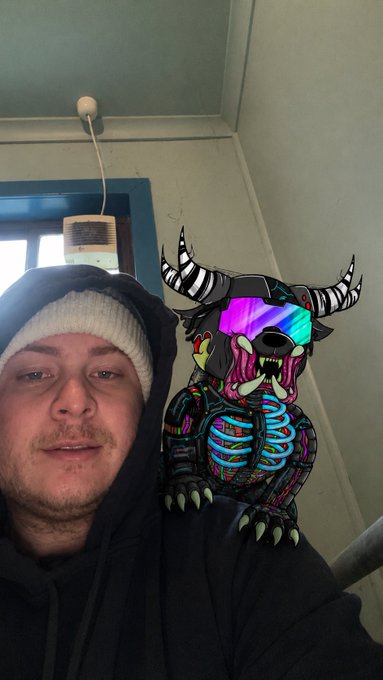 A man wearing a beanie and hoodie takes a selfie, with a colorful, neon-accented digital illustration of a horned mutant creature perched on his shoulder. The vibrant, cartoonish character—featuring skull-like facial features, rainbow-hued bones, and striped horns—adds a playful, monster-themed touch that fits the "Mutant Monday" vibe of the post.