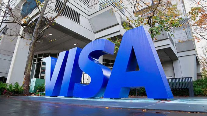A large blue 3D 'VISA' sign displayed outside a modern building, with trees showing autumn foliage in the background.