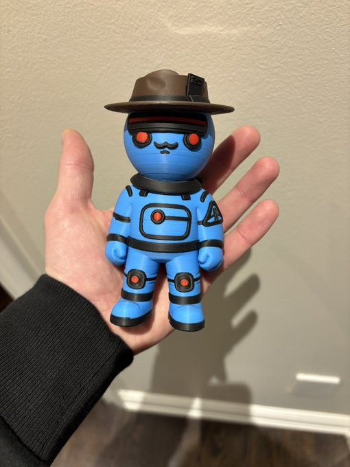 A hand proudly displays a 3D-printed blue figurine from the SpaceRidersXYZ NFT collection, featuring a small character with a mustache, red glowing eyes, and a brown fedora hat, gifted by @enchanted_nfts.