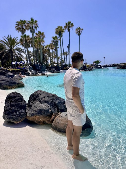 The image shows a man standing near some large rocks by a body of water, possibly an ocean or a pool. The man appears to be wearing shorts and sunglasses as he looks out at the water. There are several palm trees visible nearby, adding to the tropical atmosphere. In the background, there is a beach area with people enjoying their time in the sun. A post on social media features a prize pool of 10,000 $APE for those who reach the top of the leaderboard and mentions that there's also a random NFT available for participation.
