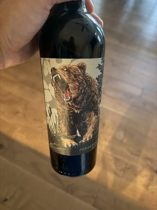 A hand holds up a wine bottle with a label featuring a roaring bear illustration and the text 'JUGGERNAUT' being held over a wooden floor.