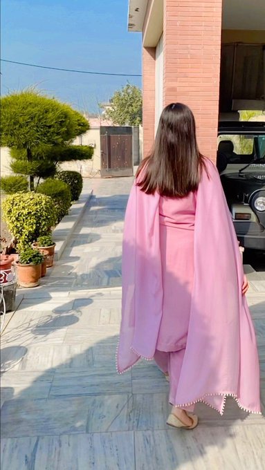 A woman with long dark hair, seen from behind, walks across a marble driveway wearing a flowing pink shalwar kameez outfit.