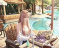 A young woman with long hair sits at a table by a swimming pool wearing a white top, holding a menu with a plate of food and a drink on the table.