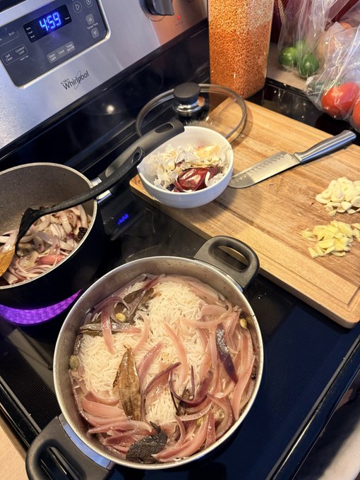 A pot of rice with onions and anchovies cooks on a stove alongside a cutting board with chopped garlic and ginger, near a container of red lentils.