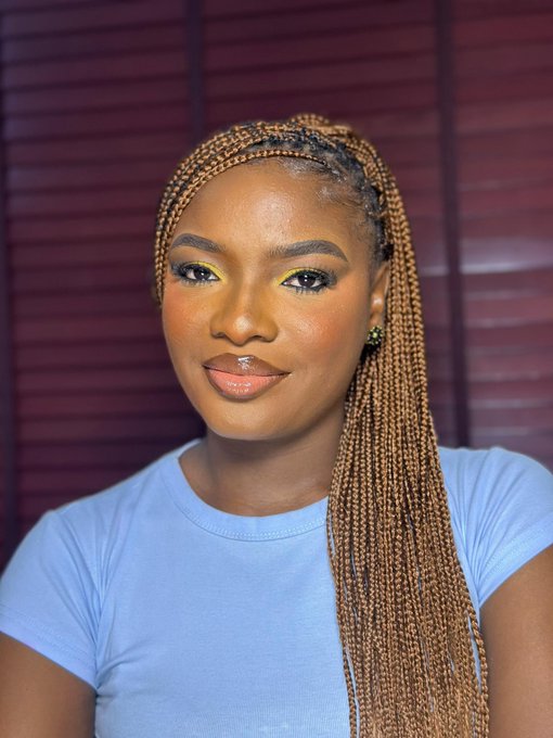 A portrait of a smiling woman with long blonde braided hair, wearing a light blue top and yellow eyeshadow.