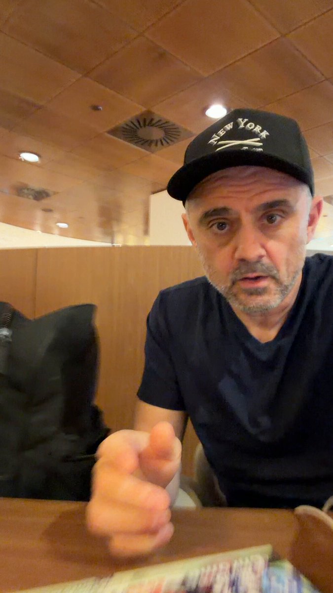 The image shows a man wearing a black baseball cap sitting at a table, pointing his finger towards something or someone outside of the frame. The background features wooden walls with a ceiling vent, suggesting that they are likely inside an establishment such as a restaurant or cafe. There is also a book visible on the table next to the man's chair.