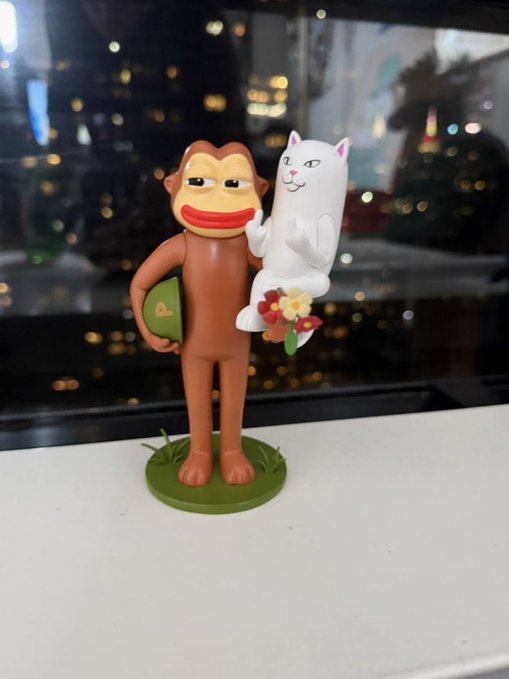 A brown monkey figurine holds a white cat and flowers on a green base, displayed on a windowsill with a city skyline at night in the background. The collectible-style sculpture visually fits the crypto-art theme suggested by the "Minted on @base" caption, as it resembles a quirky NFT-style character.