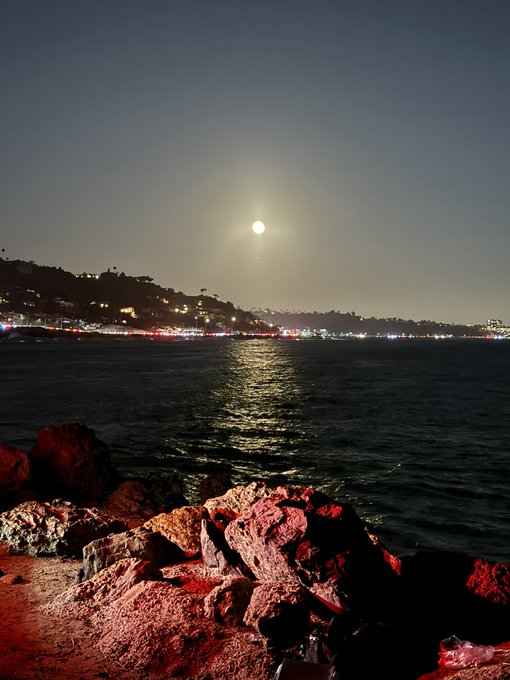 A nighttime ocean scene features a full moon reflecting on the water, with a distant illuminated coastline and rocky shore in the foreground illuminated by red light.