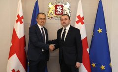 The image shows two men shaking hands over flags representing the European Union (EU) and the Georgian national flag, symbolizing a diplomatic agreement or collaboration between the two countries. The man on the left side of the photo is wearing a suit and tie, while the other man on the right side has his arms crossed in front of him. Both men are smiling as they engage in this handshake, which signifies a positive relationship or cooperation between their respective nations.