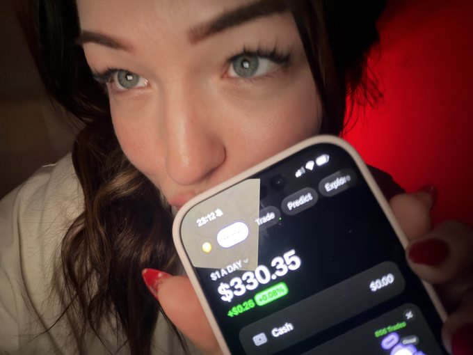 A close-up selfie of a woman holding up her smartphone displaying a stock trading app showing 'STADY' at $330.35, with an increase of +$8.36 (+2.58%).